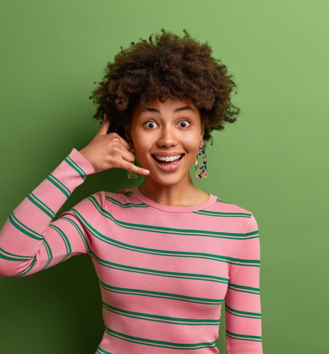 Positive dark skinned curly woman makes call me gesture keeps hands shaped like phone wears striped jumper poses against green background. Cheerful ethnic lady asks for your telephone number